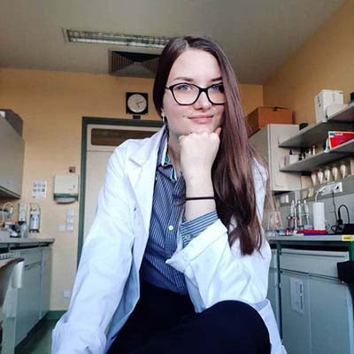 Young woman in a lab coat, sitting in a laboratory, rests her chin on her hand. She appears confident and thoughtful, surrounded by lab equipment.